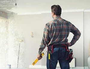 Rear view of a handyman in a tool belt holding a level and looking at a wall under renovation.