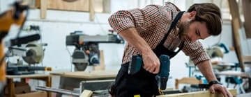 Professional carpenter using a cordless drill to work on a wooden project in a workshop