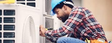 Technician in a blue hard hat and plaid shirt repairing an outdoor air conditioning unit