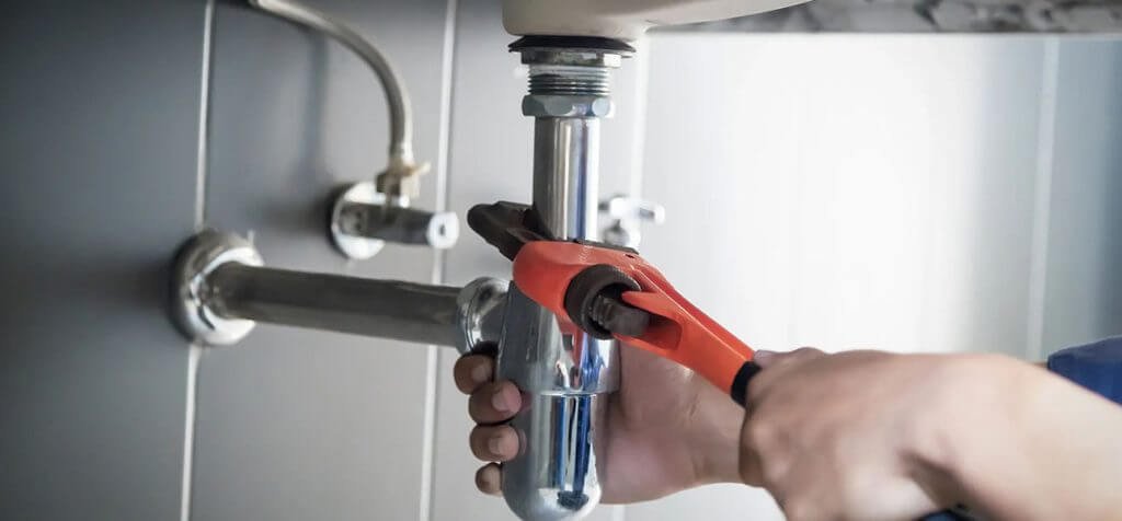 Hands using a large orange pipe wrench to tighten a chrome sink drain pipe