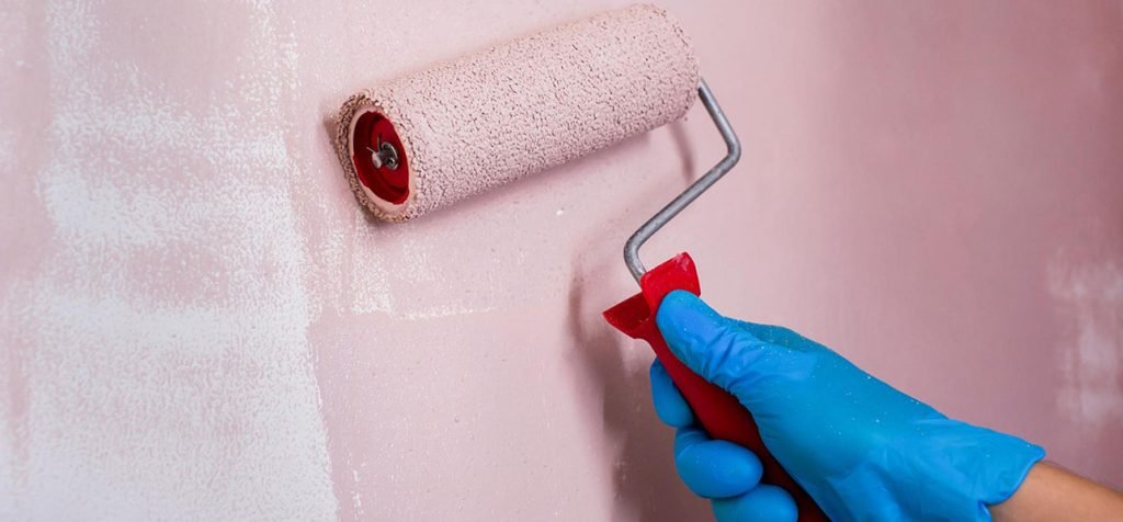 A hand wearing a blue glove paints the wall pink with a roller
