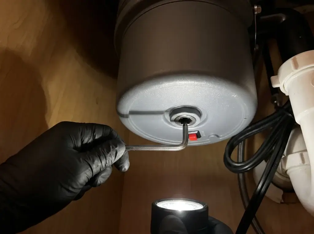 Hex key in the bottom socket of a jammed garbage disposal next to the red reset button